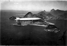 Una fotografía en blanco y negro.  El Graf Zeppelin vuela de izquierda a derecha, con un sol alto que se refleja en su envoltura.  Las dos góndolas del motor derecho y trasero son visibles, y la base misma de la sala de control está atrapando el sol.  Detrás hay rocas, montañas y mar y una gran ciudad, Río de Janeiro, construida alrededor de una bahía.