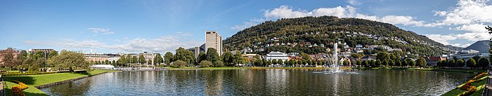 Un lago urbano con un chorro de agua en el medio, una colina redonda detrás, algunos árboles y edificios alrededor.