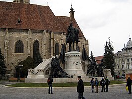 Het Matthias Corvinus-monument vóór de Sint-Michielskerk op de Piața Unirii. Uiterst rechts staat een Status-gebouw op de hoek met de Strada Iuliu Maniu.