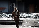 A man runs out from the cover of a destroyed building, its shattered glass lying in a frozen wave of glittering shards at its base.