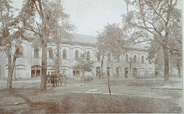 Black and white photograph of a long building, with trees and a group of soldiers