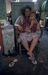 An adult woman sits while nursing a boy toddler who sits in a sling across her body.