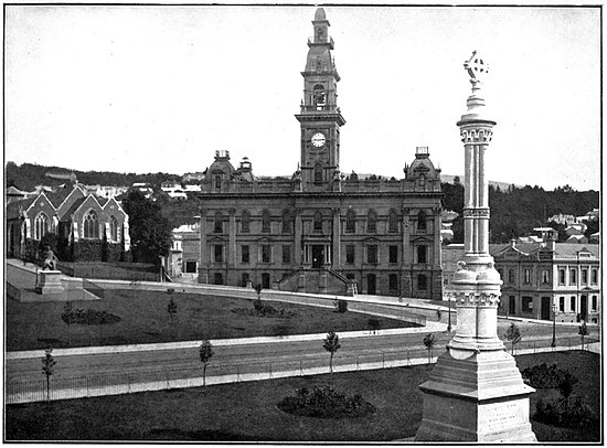 The city centre of Dunedin. On the left of the photo is a church, the size of which is dwarfed by the larger building in the middle of the photo, which has a clock face on the tower. There is a statue in the foreground of the photo that has a cross inside a circle at the top.