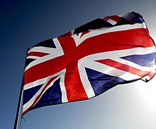 Union Flag flies on a flagpole with clear sky and sun behind.