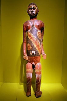 Изображение коричневой деревянной стоящей мужской фигуры, частично окрашенной крупным черным, желтым и белым пигментом, фигура находится в выставочном шкафу на зеленом фоне.