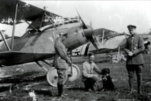 Photo noir et blanc de trois hommes en uniforme, dont l'un est agenouillé près d'un chien. Derrière eux, on peut voir des avions militaires de la Première Guerre mondiale.