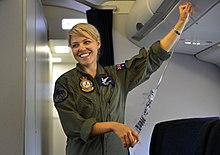 Woman in olive-green fatigues holding passenger oxygen mask in aircraft cabin