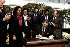 Fotografija na kojoj se nalaze Coretta Scott King koja stoji pokraj Ronalda Reagana koji sjedi i potpisuje zakon. Oko njih se nalazi šestero drugih ljudi.