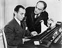 Two men sitting behind a piano, writing music.