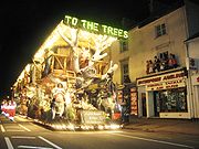 A large vehicle lit by many lights and carrying people dressed in costume in a darkened street. People stand on the balconies of the shops behind.