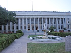 Vue partielle de la façade d'un bâtiment blanc à colonnes. Au premier plan sont visibles des massifs et une fontaine avec une statue de cupidon.