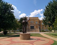 A statue of a woman in Native American costume holding a flat drum and striking it with a mallet. In the background is a brick building.