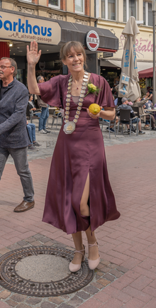 Das Foto zeigt Eva Döhla nach rechts blickend und winkend. In ihrer linken Hand hält sie eine Zitrone. Sie trägt ein weinrotes Kleid.