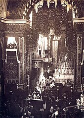 Looking down upon an assembly in a large, vaulted cathedral with a figure sitting on a large, canopied throne to the left of an altar