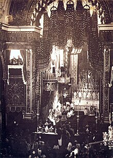 Blick auf eine Versammlung in einer großen, gewölbten Kathedrale mit einer Figur, die auf einem großen Thron mit Baldachin links von einem Altar sitzt