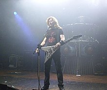 Muž s dlouhými zrzavými vlasy stojící na pódiu před mikrofonem