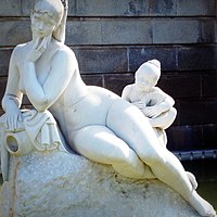 Escultura de piedra blanca que representa a una mujer sentada, recostada ligeramente sobre su lado derecho y una niña sentada. Ambas desnudastras ella