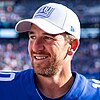 Headshot of Eli Manning smilining, in uniform and wearing a New York Giants hat