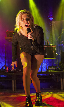 A woman singing into a microphone, wearing black clothing. In the background, drums and turntables can be seen.