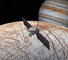La sonde occupe le centre de l'image, composée notamment d'une parabole et de deux grands panneaux solaires. Sous elle, la surface grise et brune d'Europe avec ses lineae. Derrière, Jupiter.