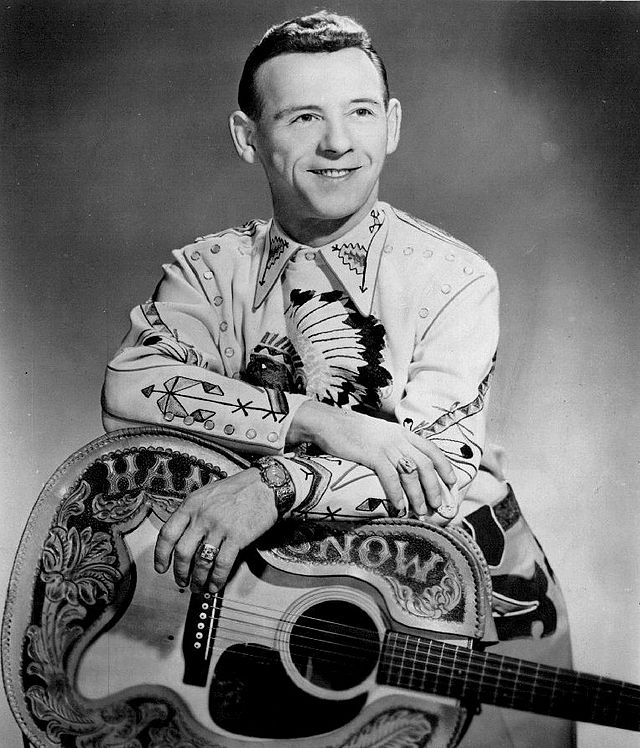 A man wearing an elaborately patterned shirt, leaning on a guitar