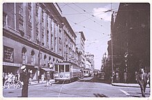 Black and white photo of a downtown intersection, with a streetcar along the road and pedestrians along the sidewalks