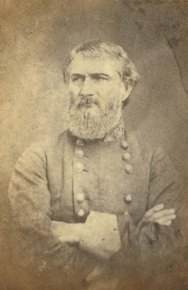 Sepia tone photo of a frowning, bearded man wearing a gray military uniform. His arms are folded.