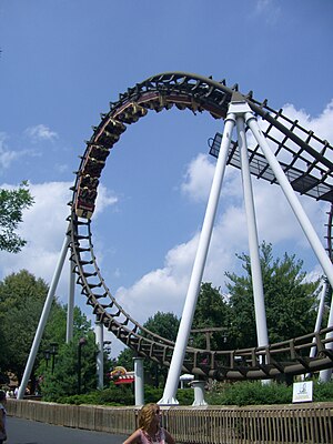 A train is seen inverted in the latter half of the cobra roll element when it was known as the Sidewinder. The roller coaster's brown track and white supports are prominent among the greenery in the background.