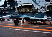 Aqua and blue jet aircraft on aircraft carrier deck, with a group of men standing close-by