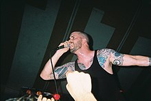 Vocalist Andrew Schwab performs at a concert. He has shaved his head and is wearing black sleeveless shirt that displays his tattoos.