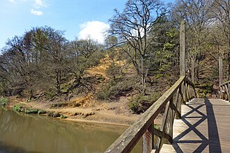 This sandy bank has been exposed by the River Wey as it cuts through the Upper Greensand stratum (mid Cretaceous) near Shalford The North Downs Way crossing the River Wey near Shalford in Surrey (geograph 5744682).jpg