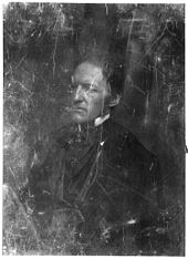 A scratched, half-length photographic portrait of a middle aged man in formal mid-19th century dress, facing left