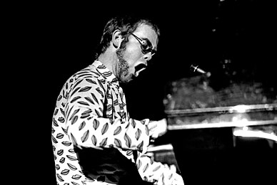 A black and white photo of Elton John in an elaborate glam costume, playing the piano.
