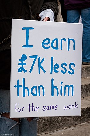 A woman holding up a sign protesting that she earns less than a "him" (a male coworker) for the same work