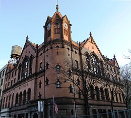 Photographie d'un bâtiment en brique avec pignons, arcades, tour d'angle octogonale et horloge à quatre faces.