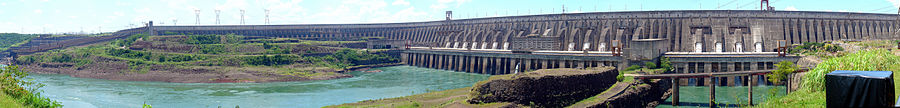 Uma grande barragem de concreto cinzenta, com diversas comportas, se estende horizontalmente. Dela saem dois fluxos de água calma. Ao fundo, algumas torres de transmissão de energia.