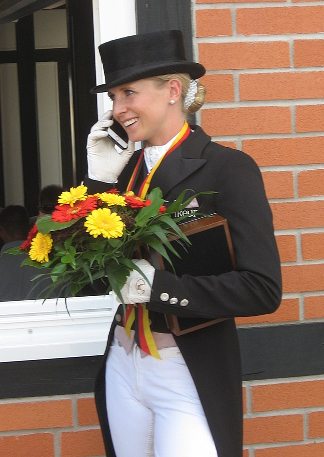 freundliche Dame mit schwarzem Hut, weißen Hosen, Frack und Blumen in der Hand bei einer Reit-Meisterschaft telefoniert