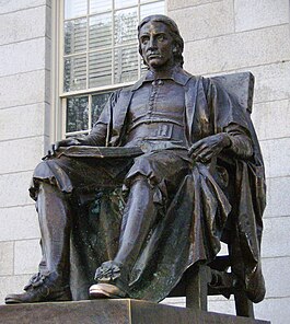 Eine Bronzeskulptur auf einem hohen Granitsockel eines Mannes, der mit einem aufgeschlagenen Buch auf dem Schoß auf einem Stuhl sitzt.Die Statue als Ganzes ist dunkel verwittert, aber die Spitze des linken Schuhs der Figur glänzt wie von häufigem Reiben.