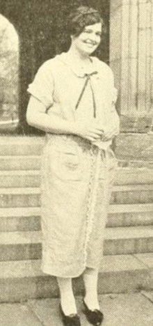 A young white woman with dark hair, standing in front of steps and smiling, wearing a casual dress with pockets and a ribbon tie at the collar