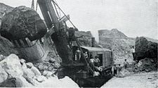 Черно-белая фотография, показывающая часть другой, похожей паровой лопаты на участке нарушенной земли с большим камнем в ведре.