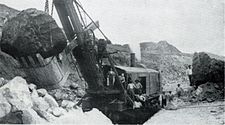 Ein Schwarzweißfoto, das einen Teil einer anderen, ähnlichen Dampfschaufel in einem Gebiet gestörter Erde mit einem großen Stein im Eimer zeigt