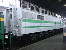 Foto colorida mostrando a traseira de uma locomotiva branca com uma listra verde.  Uma porta permite o movimento com o trem, mas não há janela da cabine.
