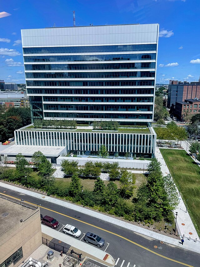 A picture of glass skyscraper with a white facade that is 10-15 stories tall, taken from another nearby tower.