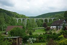 Kamenný víceobloukový viadukt přes údolí v obci Novina. V pozadí se nachází Ještědský hřbet (samotný nejvyšší vrchol však na tomto záběru není vidět). V blízkosti viaduktu jsou k vidění tradiční hrázděné domy a usedlosti. Snímek pochází z letního období a celá krajina je plná bující zeleně.