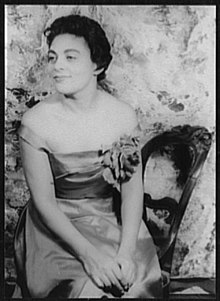 An African-American woman, seated, wearing a gown with a low neckline and bare shoulders; she is smiling, with her face turned to her right