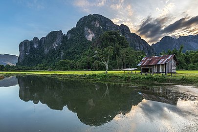 Water reflection of karst mountains, wooden hut, trees and colorful clouds at sunset with green paddy fields, Vang Vieng, Laos