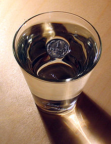 Photo of a drinking glass filled with water from above.  In the middle of the water surface there is a metal coin that is labeled "1 Pfennig".  Around the coin you can see that the surface of the water is dented where it touches the coin.