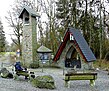 Eine kleine steinerne Kapelle im Wald, die ein mit schwarzen Ziegeln gedecktes Spitzdach hat. Neben der Kapelle steht ein Glockenturm, der sie überragt, vor der Kapelle stehen zwei Sitzbänke.