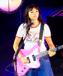 Beabadoobee onstage playing an electric pastel pink guitar while wearing a manga t-shirt.