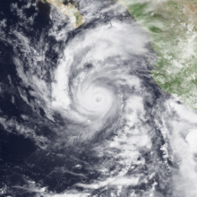 A hurricane over navy blue water with beige and green pieces of land to the top and to the right; the hurricane's cloud pattern resembles the number 6, with a cloudy eye in the middle; some streaky but deep clouds in the lower right are those of a nearby tropical storm